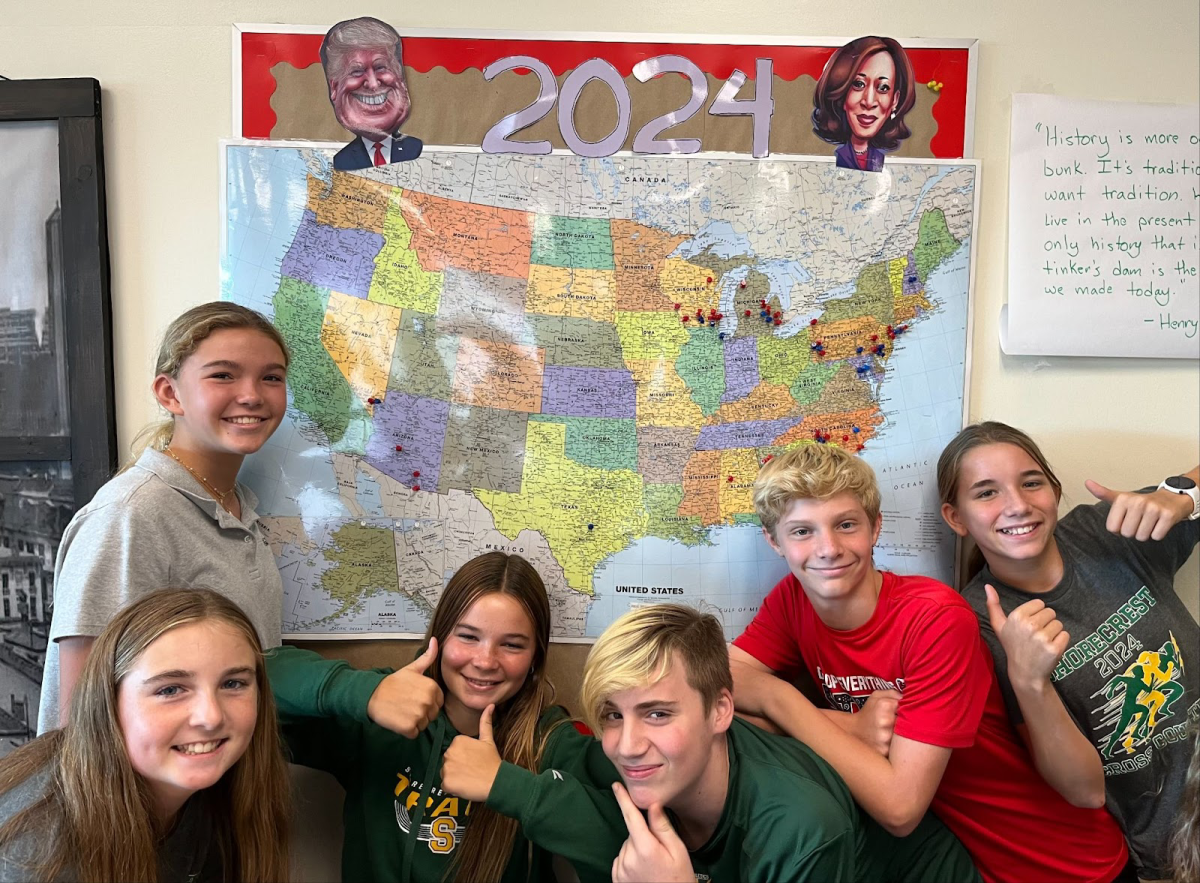 7th graders (From left to right: Georgia S., Kai E., Sophia S., Frank C., John J., and Alessandra N. ) pose in front of an election-themed bulletin board and map. Hodgson repeats this election-themed unit every four years during a presidential cycle. Photo by David Hodgson