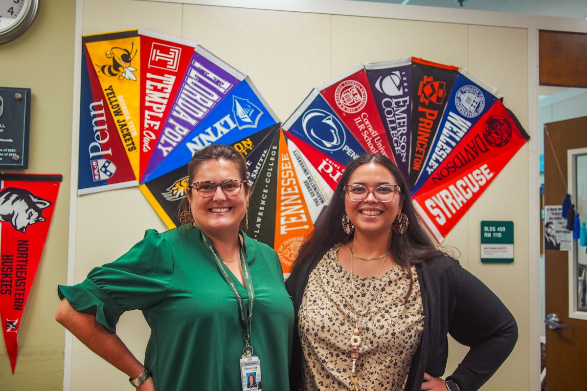 Senior Associate Director of College Counseling Meghan Schneider smiles with Assistant Director of College Counseling Victoria Scott in front of their plethora of college flags displayed on their office wall. Students rely on the help of counselors to manage the college admissions process smoothly.

