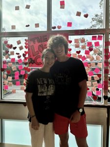 Junior couple Luna Gillis and Charlie Cowan pose for a photo in front of "The Love Club" sign. Having met in fourth grade, they have grown close over the years, forming strong connections with each other's families. Their relationship challenges many common Gen Z stereotypes.